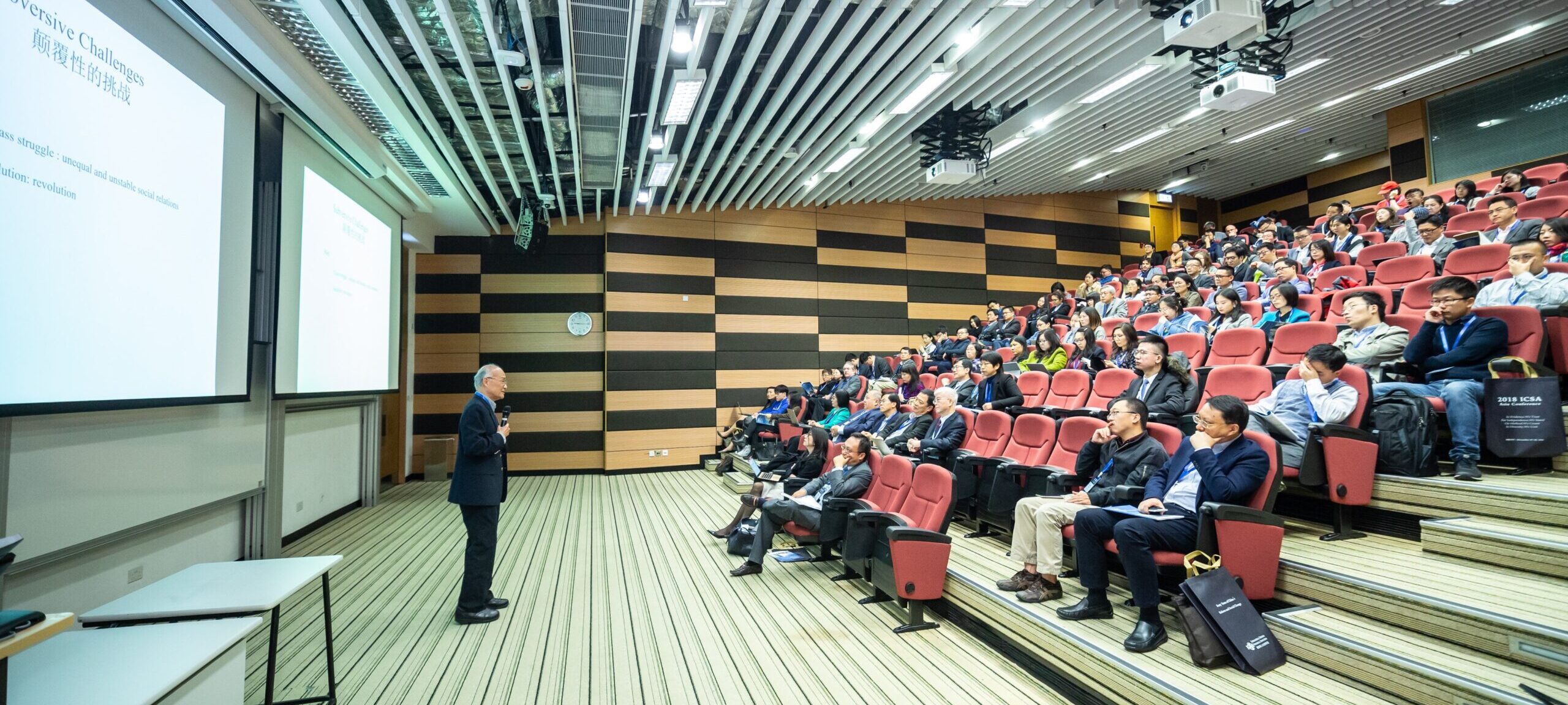 Man speaking in front of a slideshow to an audience sitting in a lecture hall.