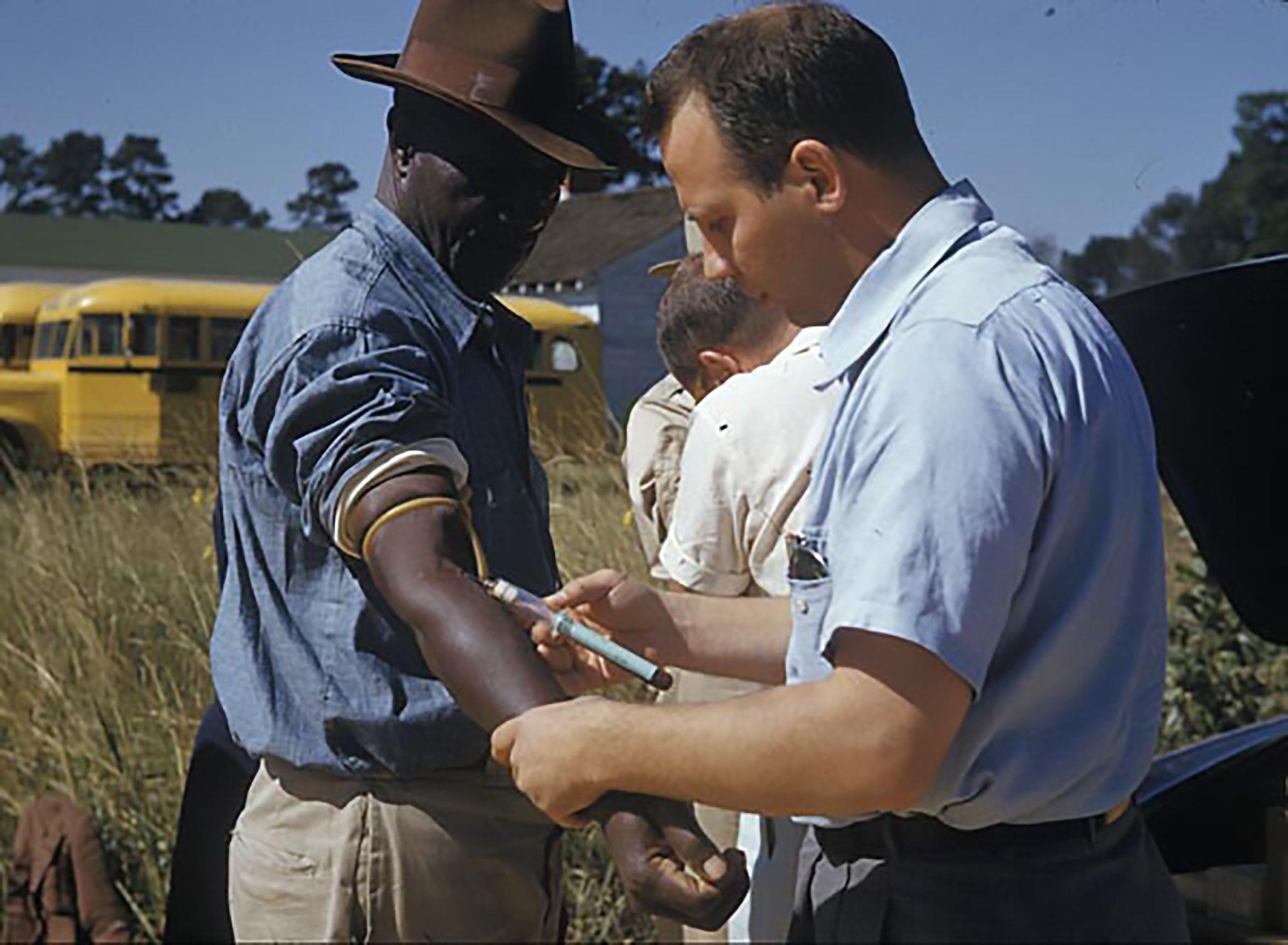 A white man administers an injection to an African American man standing outside in a field.