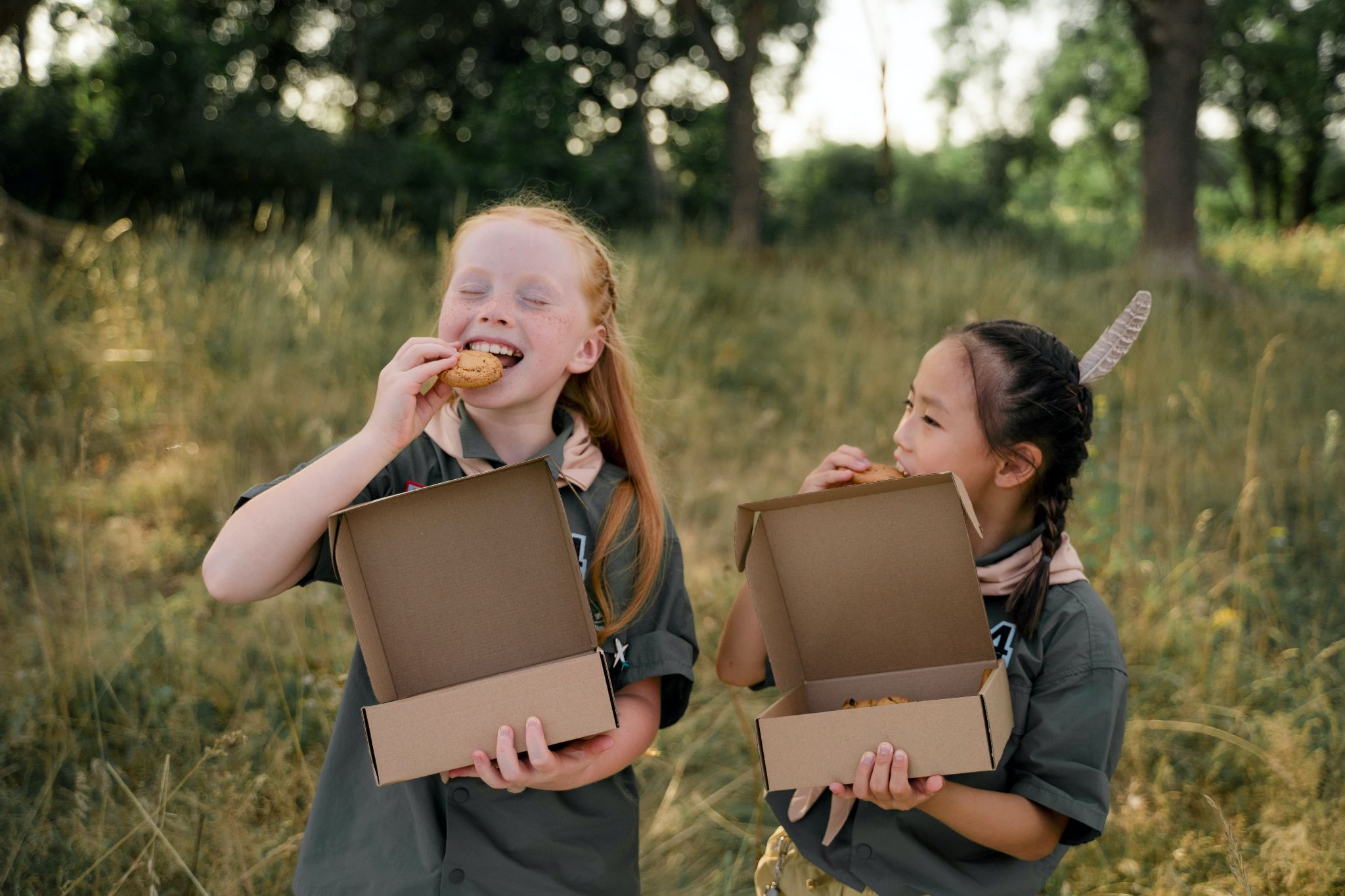 Two girls in scout uniforms eating cookies from boxes each are holding.