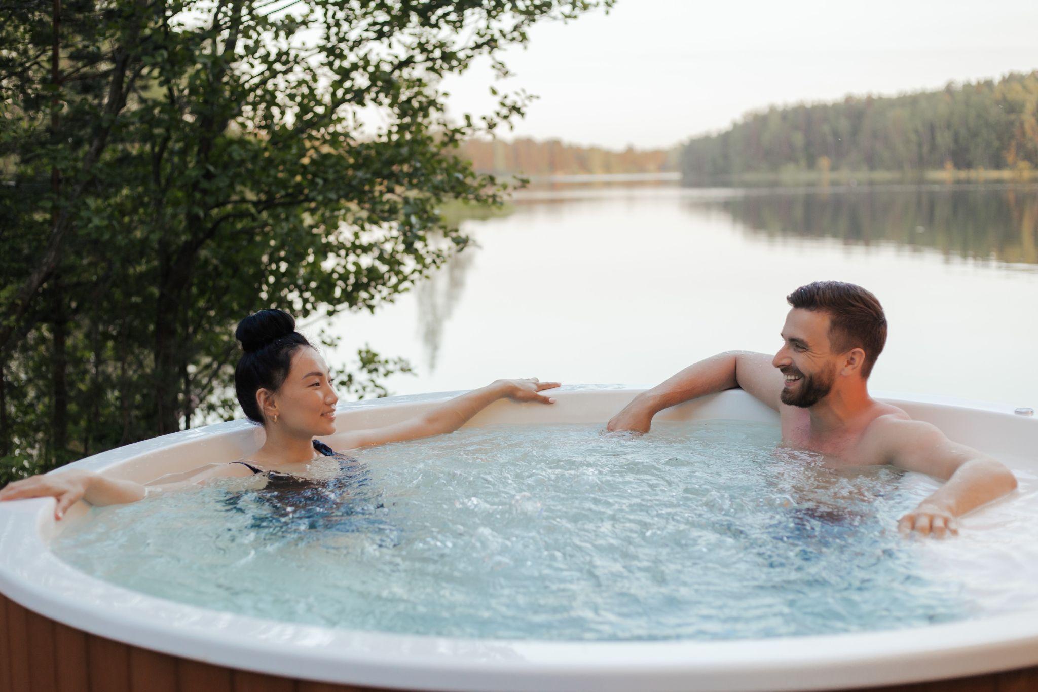 Two people lounging in an outdoor hot tub next to a wooded lake.