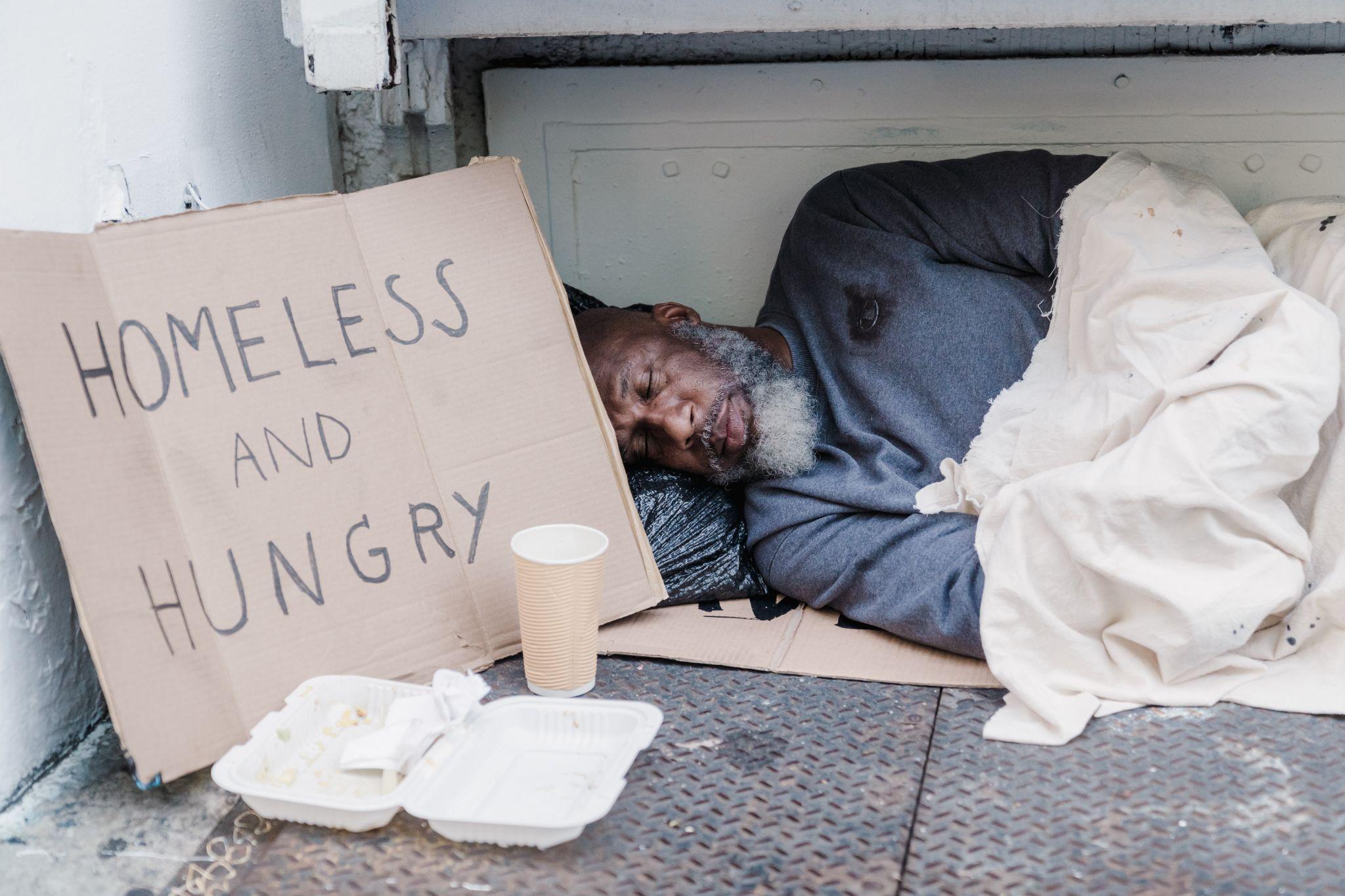 Man sleeping on the sidewalk next to a cardboard sign that reads “homeless and hungry.”
