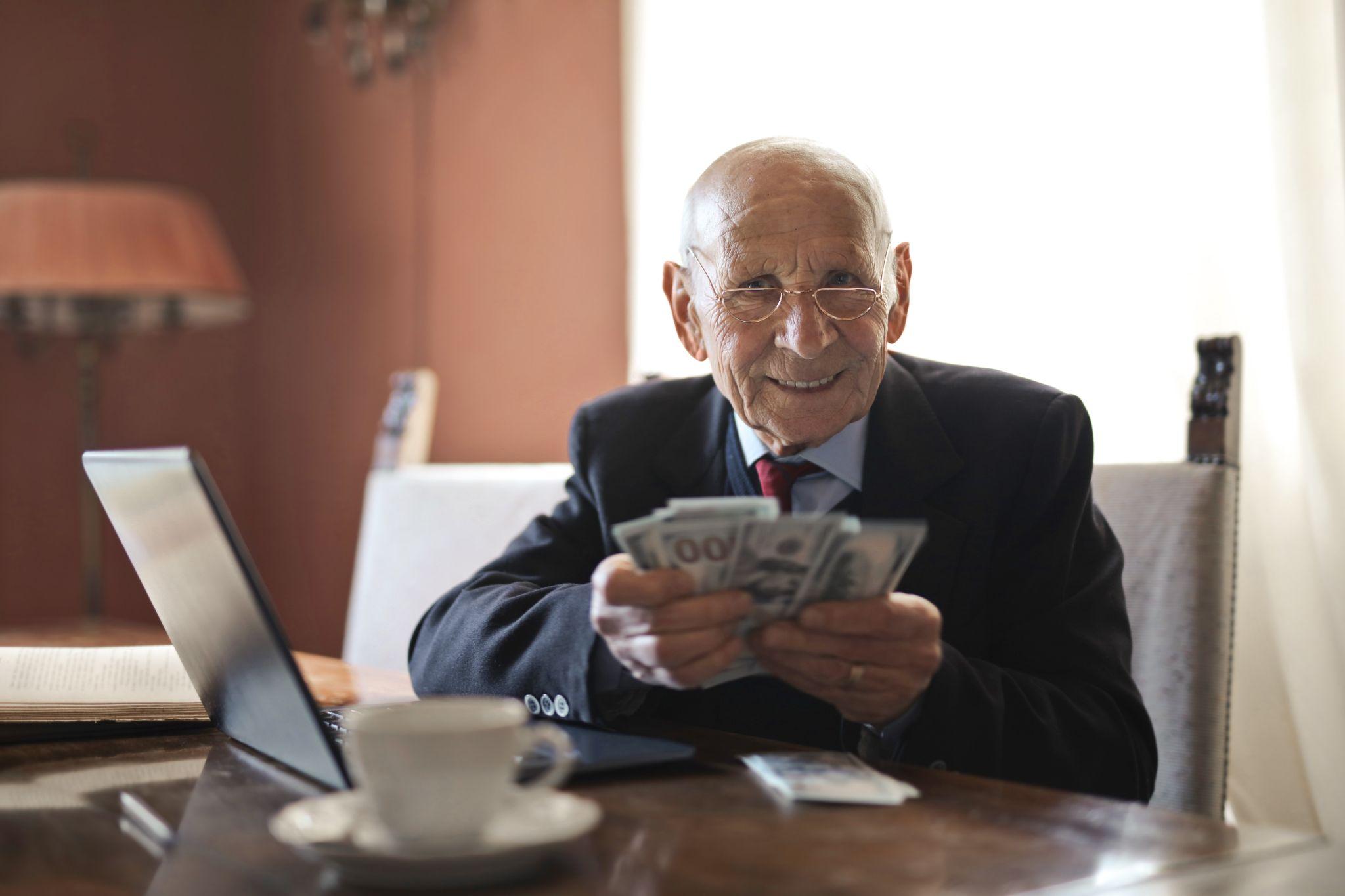 Old rich man in a suit holding $100 bills in his hand and smiling at an office desk.