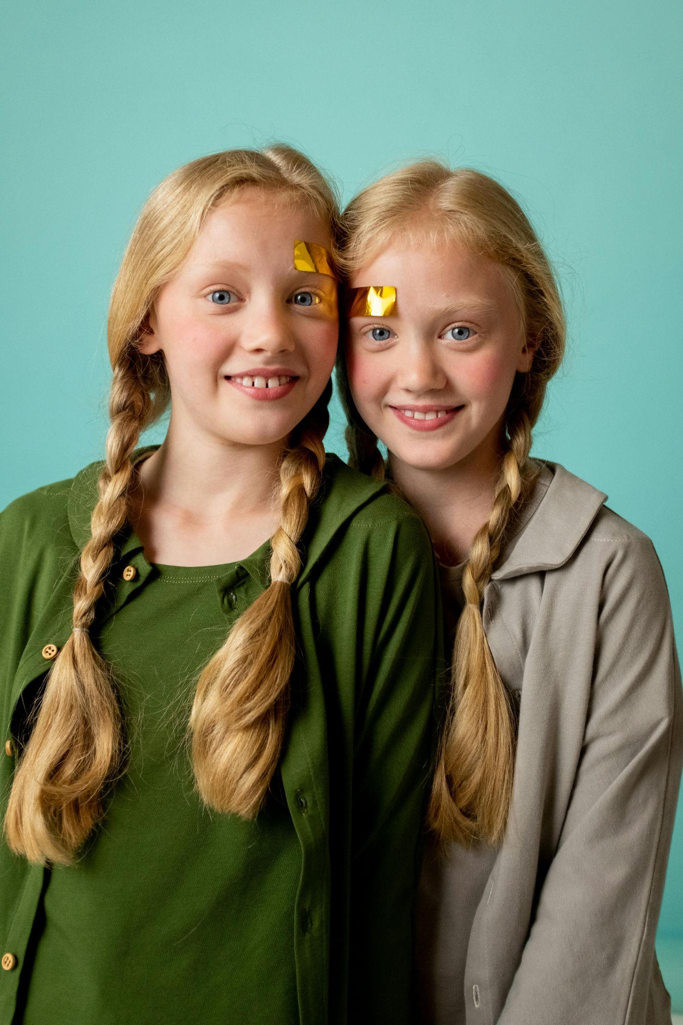 Picture of twin girls with pigtails smiling. They are largely identical, though they have shiny strips of paper stuck on different sides of their foreheads.