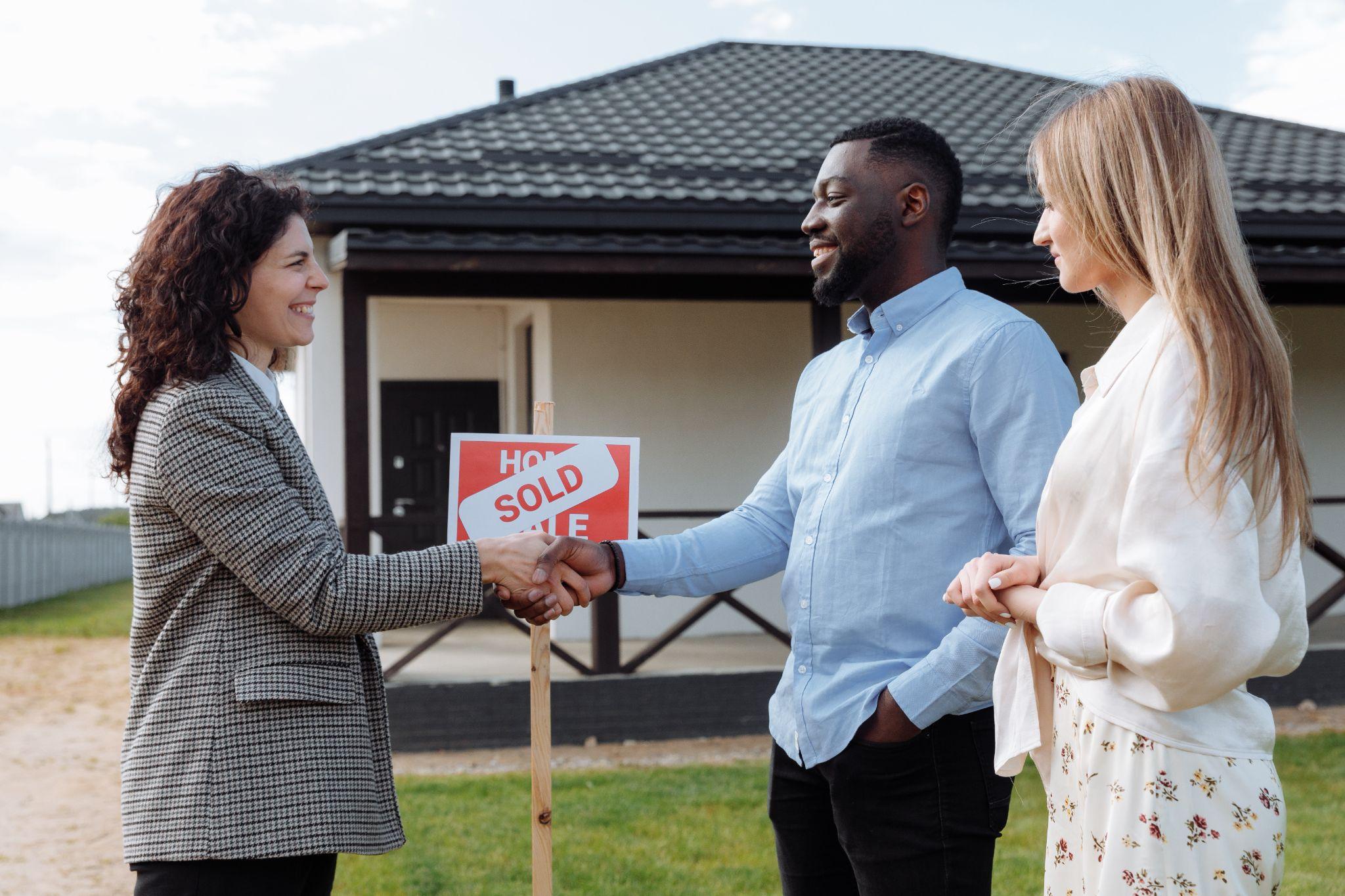 Black man shaking the hand of a real estate agent while his white partner watches in front of a house with a “Sold” sign.