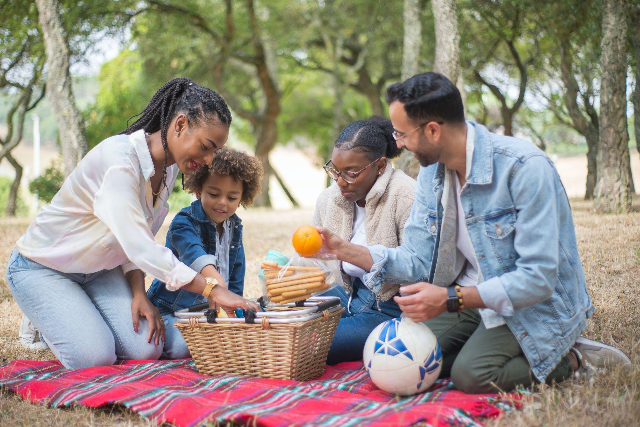 Mixed-race family sitting down on a blanket with a picnic basket in front of trees.
