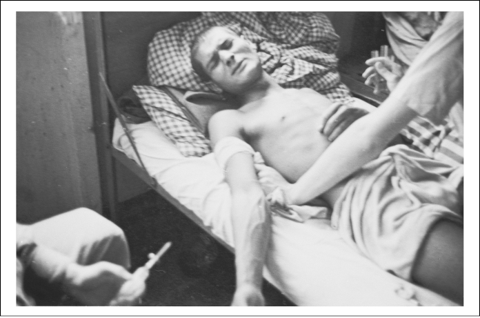 Prisoner on a bed grimacing as another person holds him and a third person holds a needle.