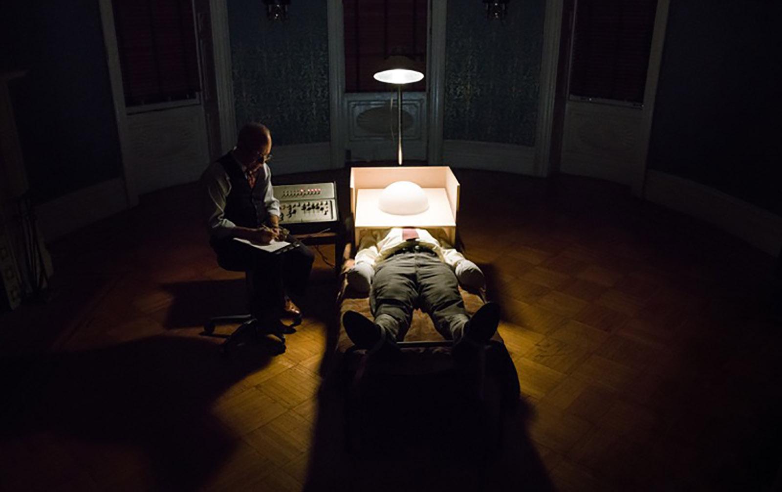 Peter Sarsgaard lying down next to a scientist with an overhead light shining on him.
