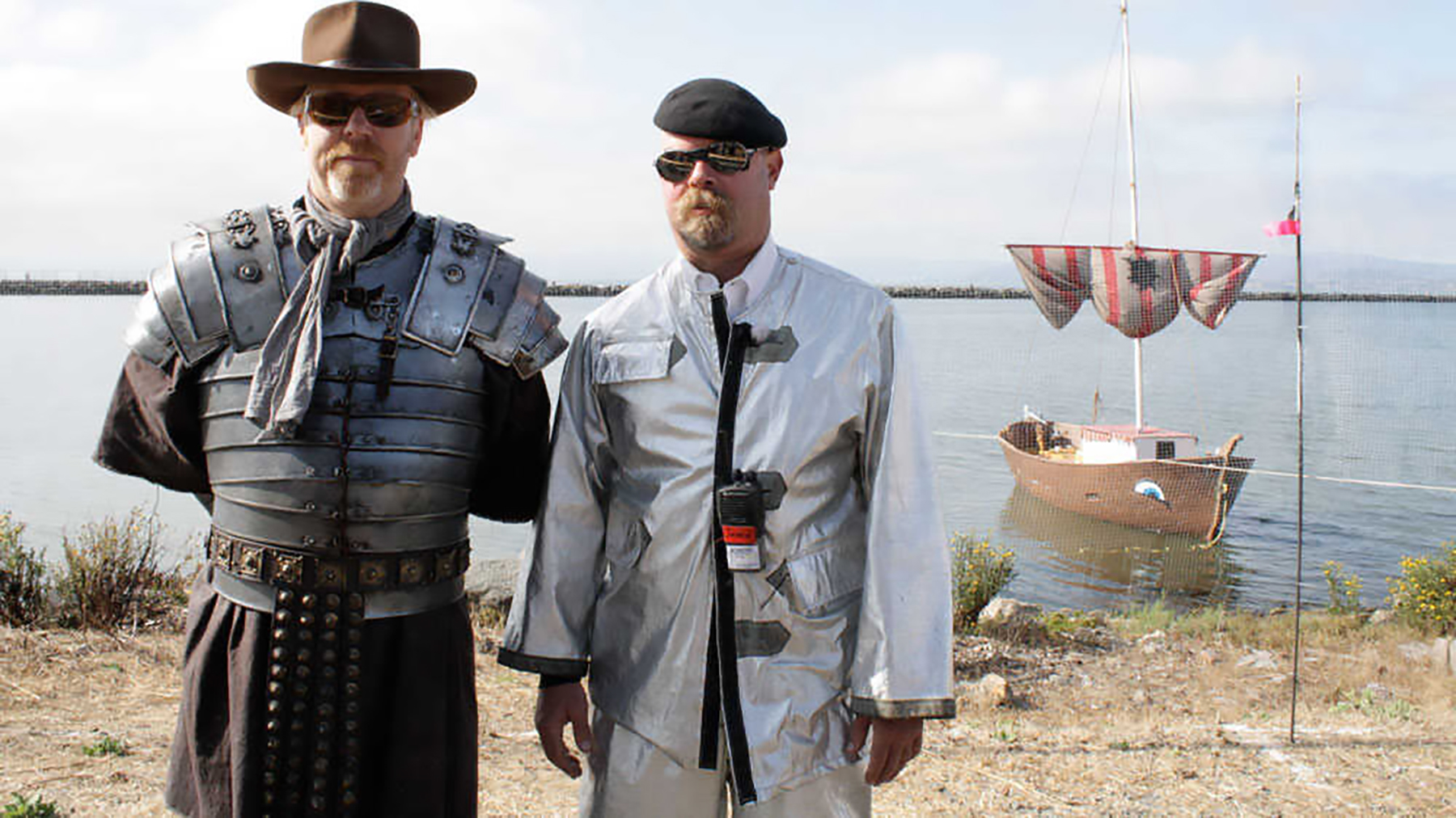 MythBusters hosts Adam Savage and Jamie Hyneman standing on a beach in front of an ancient-looking sailboat.