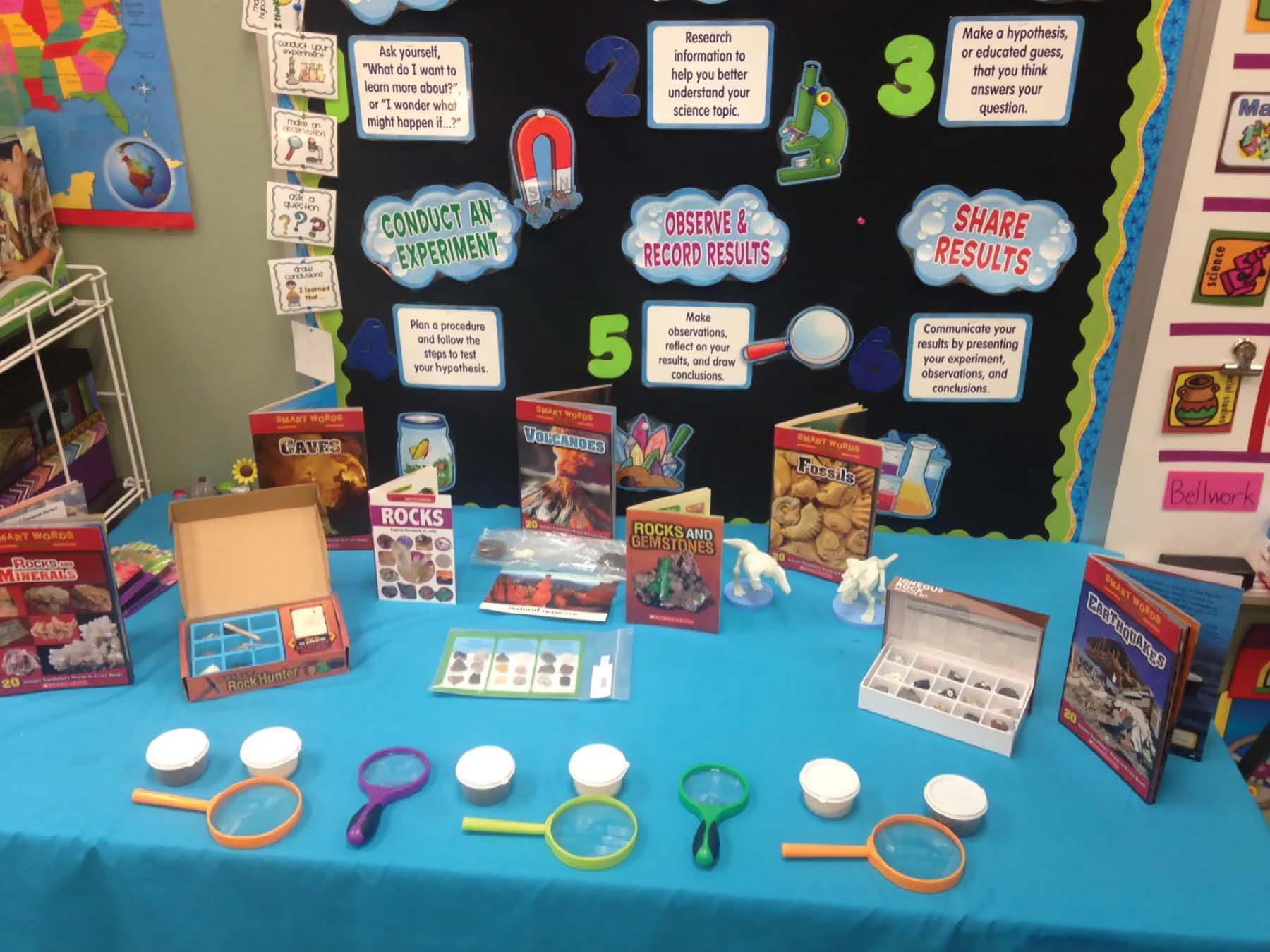 An elementary science center, with a table set up to encourage hands-on learning. On the table are science-related books, activity kits, magnifying glasses, and small containers. Behind the table is a colorful bulletin board that describes the steps of the scientific method in simple, child-friendly language.