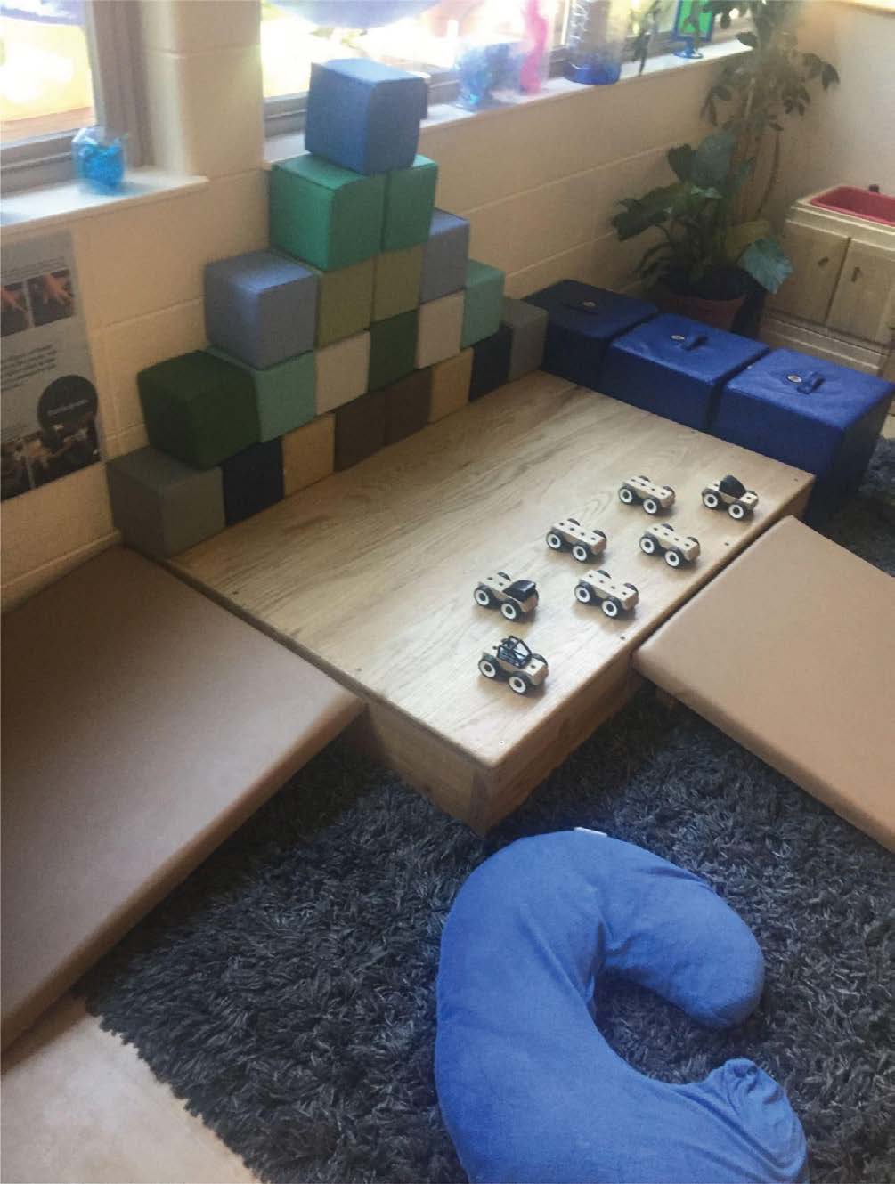 A toddler play space features a wooden platform with several small toy cars arranged on top, positioned for ramp play. Cushioned mats surround the platform for comfort and safety. Against the wall, large foam blocks are stacked in a stepped pattern, and padded seats are placed nearby. A curved blue cushion lies on a dark rug in the foreground.