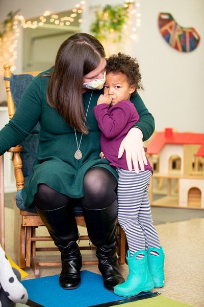 A teacher seated on a chair gently holds and leans toward a young child who stands close, resting against her. The child has a thumb near their mouth and a slightly worried expression, while the teacher's arm is wrapped protectively around them.