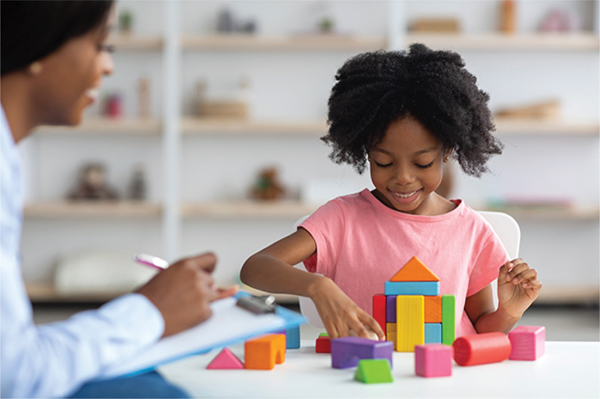 A child sits at a table, focused on arranging colorful building blocks into a small structure of various shapes, while an adult nearby holds a clipboard and takes notes, attentively observing the child's play.