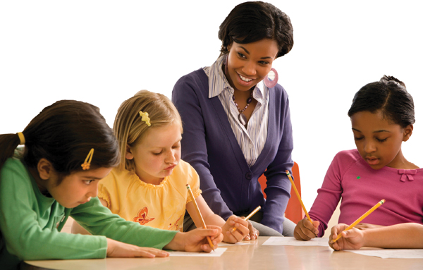 A teacher stands close to three seated children, attentively watching as they work on writing tasks. Each child holds a pencil and focuses on their individual paper.