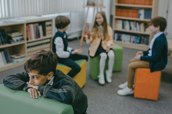 A boy sits apart from a group of three other children, resting his head on his arms and appearing withdrawn, while the others engage in conversation together.