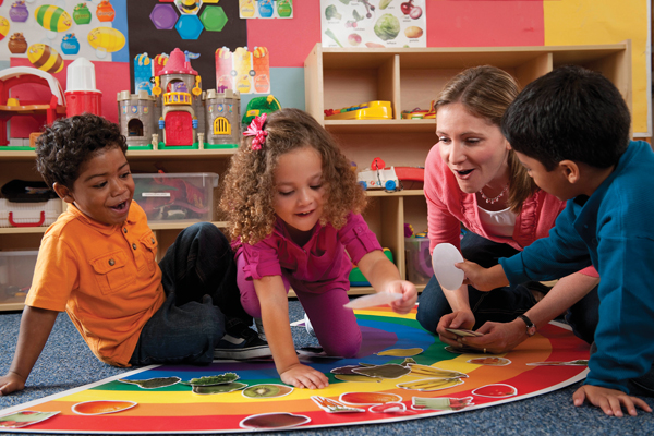 A group of young children in a bright, colorful classroom play a matching game. The children sit on the floor in front of a large poster shaped like a rainbow, matching different pictures of fruits and vegetables to the rainbow's colors. The teacher interacts with the children, guiding their play.
