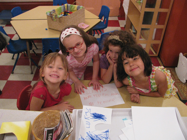 Four young children at a classroom table smile cheerfully at the camera. In front of them is a piece of paper on which they have written words in crayon together. Art supplies and additional papers are placed nearby on the table.