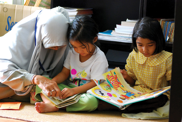 A teacher sits on the floor beside two young girls, guiding one of them through a picture book. The girl in the center listens attentively while looking at the pages, and the girl on the right reads her own colorful book.