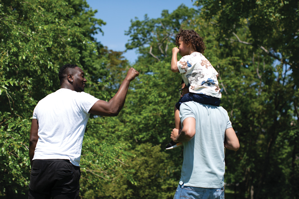A man walks beside another man carrying a child on his shoulders along a green tree-lined path. The walking man extends his fist toward the child, who leans forward to meet it.
