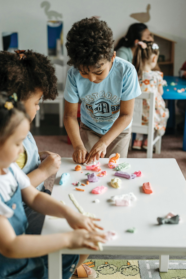 Three children are gathered around a small white table in a classroom, shaping colorful clay pieces into figures. One child stands while pressing clay, and the others sit, using their hands to mold shapes.