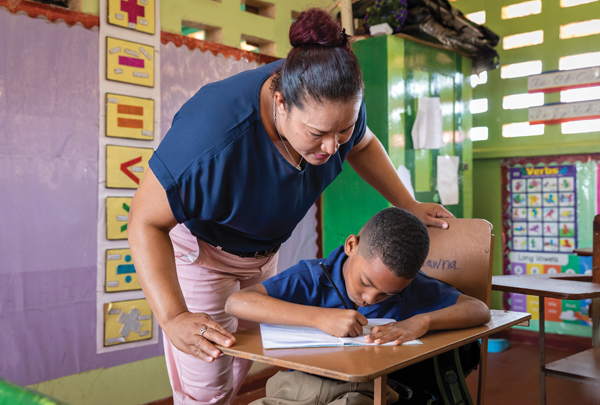 A teacher leans over a young boy seated at a desk, guiding him as he writes in a notebook.