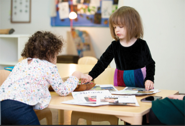 Two young children are seated at a wooden table, engaged in a collaborative activity about their families. One child leans forward, reaching toward a container while the other focuses on arranging or writing on the papers in front of her. The table is scattered with printed sheets and books.