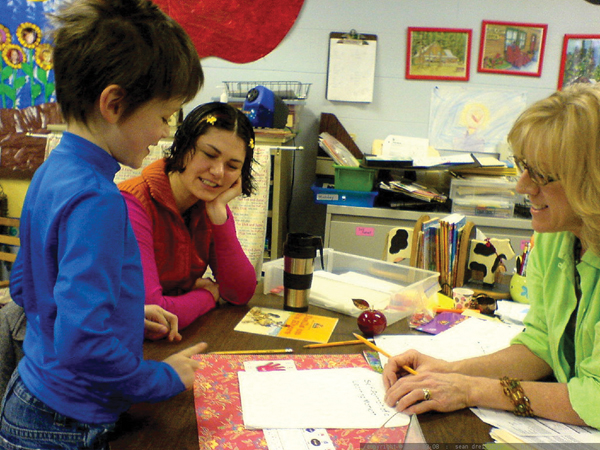 A child stands beside a table speaking with a parent and teacher in a classroom. The parent rests her hand on her face, smiling, while the teacher listens attentively, holding a pen above a sheet of paper. The table is filled with school materials such as books, folders, and stationery.