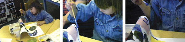 A child sits at a table painting a three-dimensional object in three sequential views. The child dips a paintbrush into green, blue, and black paints and applies them to the object with focus and careful hand movements.