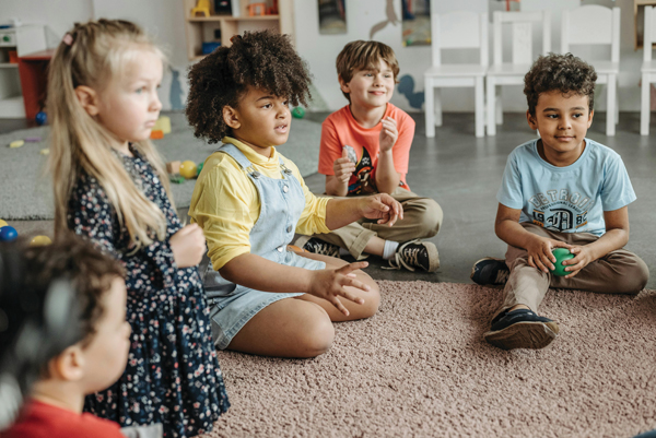 A group of young children sit together on the floor in a classroom, forming a circle. They appear engaged with one another, some holding toys and looking toward their peers.