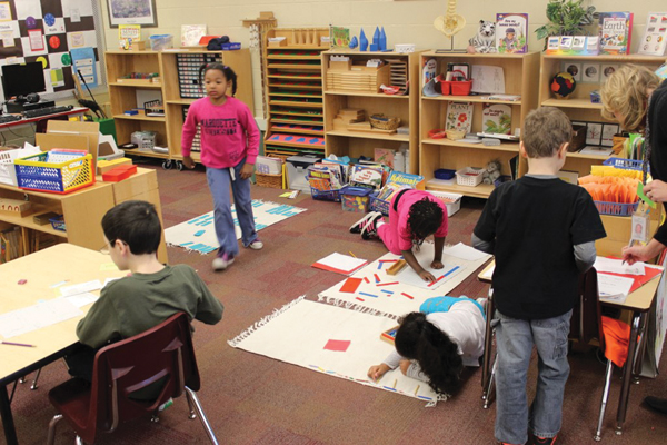 Several elementary school children are engaged in various activities in different parts of a classroom. Some children sit at tables working with paper and materials, while others work on the floor with educational tools and mats.