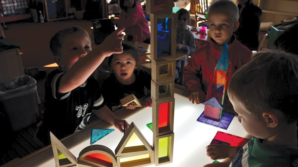 A group of preschool children are gathered around a light table, building structures with translucent, colored geometric blocks. One child is placing a piece on a tall tower, while others watch with interest.