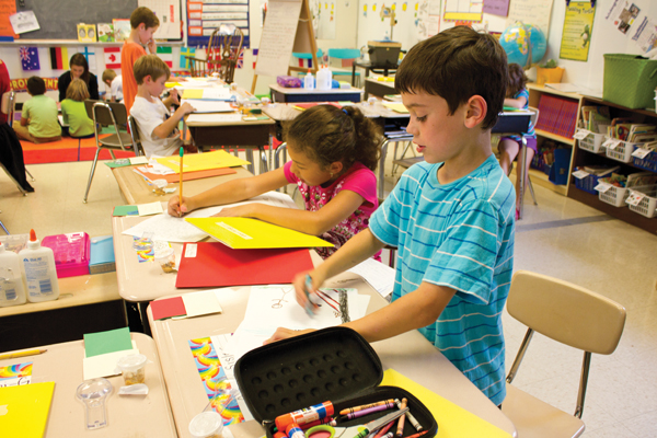 Children in a primary-grade classroom are engaged in various activities at their desks. A boy in the foreground uses glue while a girl beside him writes or draws. Supplies such as paper, folders, glue sticks, and markers are scattered on the desks.