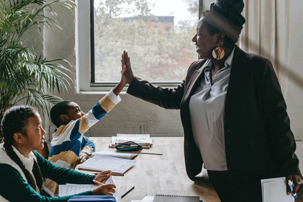 A teacher stands next to a desk and engages with a student by giving a high-five, while the student smiles and reaches up from their seat. Another student sits beside them, watching with interest. Both children have notebooks open on the desk.