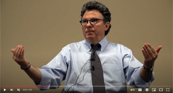 A still image from a video features Doctor Ross Greene with his arms extended outward, using a gesture that suggests he is comparing or weighing two ideas.