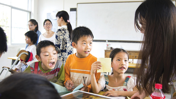 A group of children sit at desks engaged in classroom activities while a teacher leans closer to interact with them.
