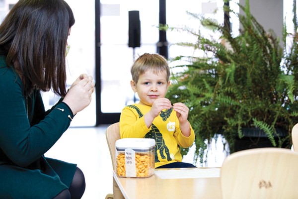 A young child sits at a table facing a teacher, making a sign language gesture while looking toward the teacher, who is responding with hand signs.