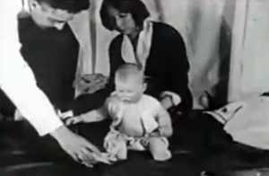 Black-and-white photograph depicting the Little Albert experiment. A young child, known as Little Albert, is seated on a mat or platform. Nearby, psychologist John Watson and a woman—possibly his assistant—are present. A white rat is placed near the child. The setting appears clinical, with minimal background distractions.