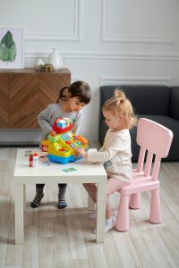 A picture of two young children engaging in symbolic play. They are at a table with a play cash register.