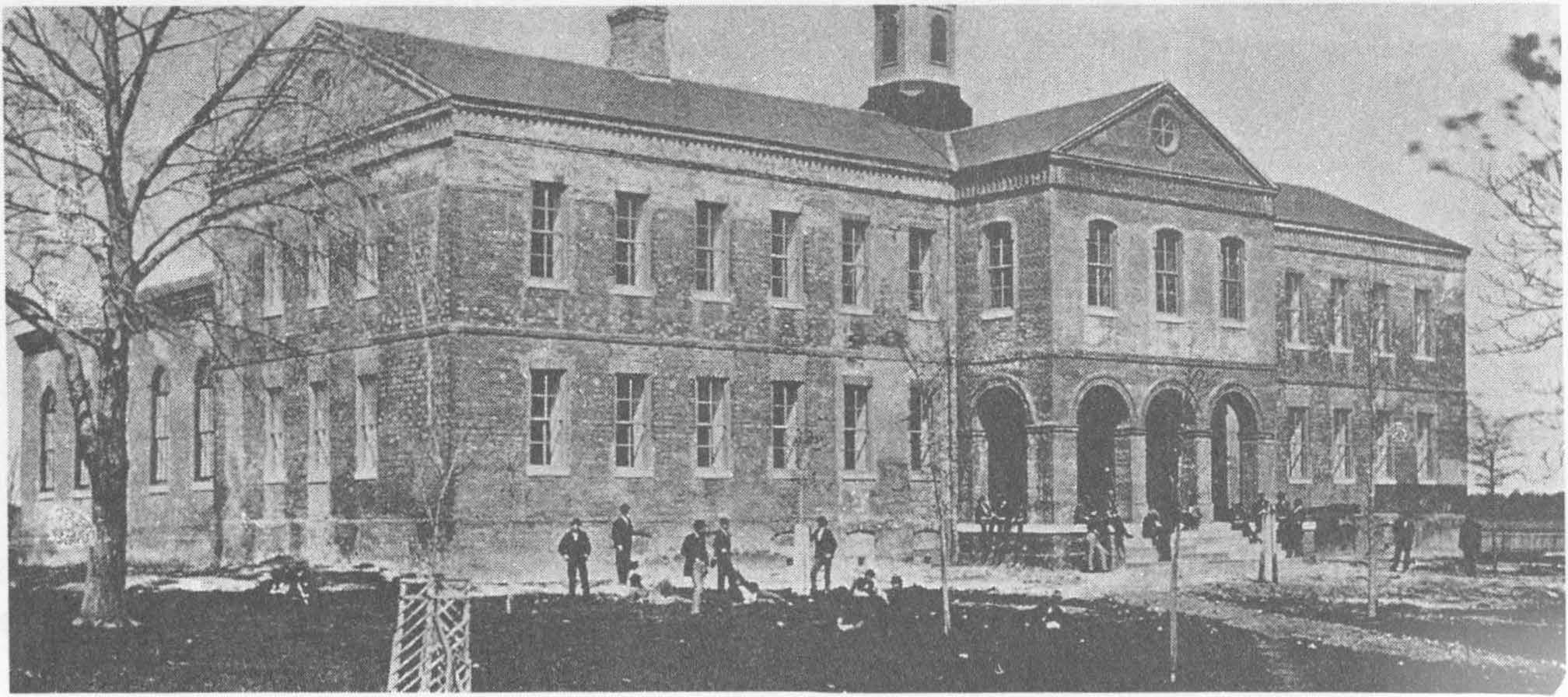 Brick building with arches and multiple windows, surrounded by people and leafless trees.