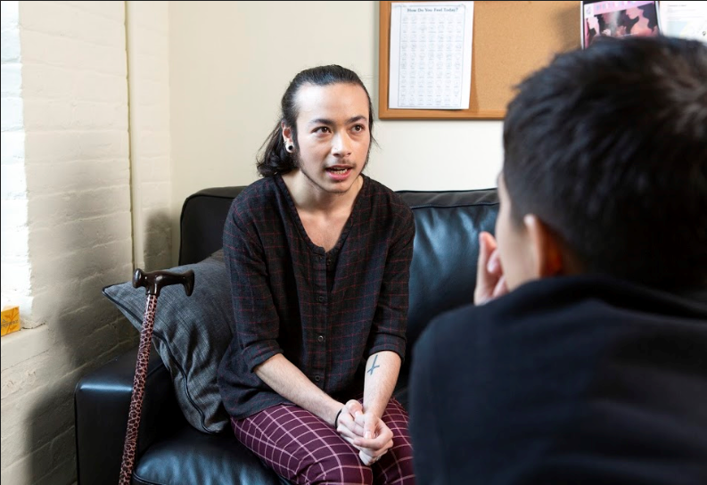 A genderqueer person sitting on a couch, talking to a therapist in a brightly-lit room