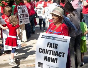 A teacher dressed in red holds a sign reading, "Fair Contract Now!" at a strike.