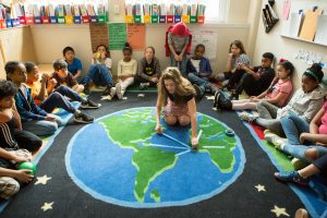 A teacher puts tape on a carpet with a globe on it as students sit at the perimeter of the carpet.