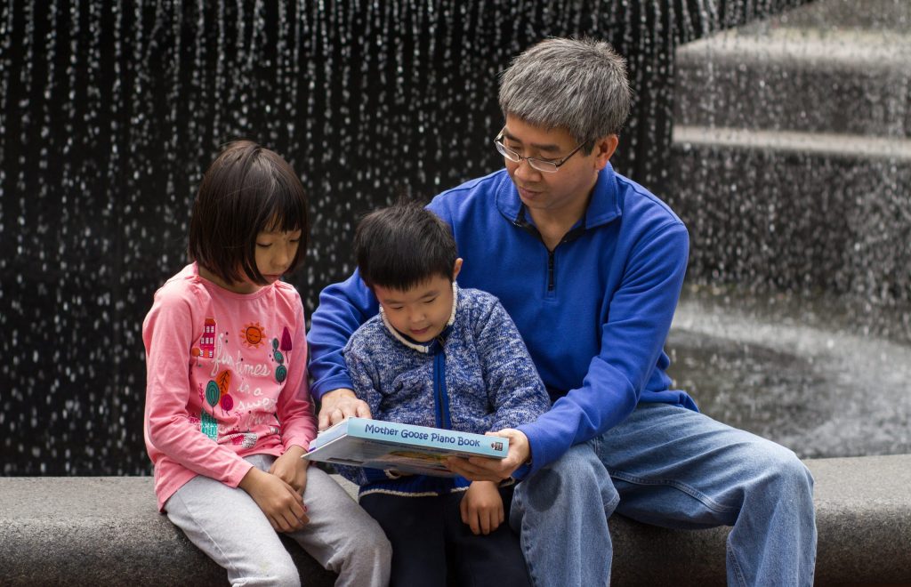 A parent and two children are sitting at a fountain. The parent holds the book in front of one of the children while the other looks on.