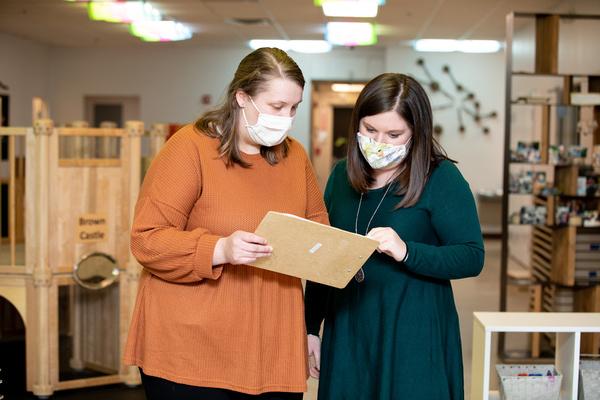 Two teachers stand in the classroom looking at a clipboard one of them is holding.