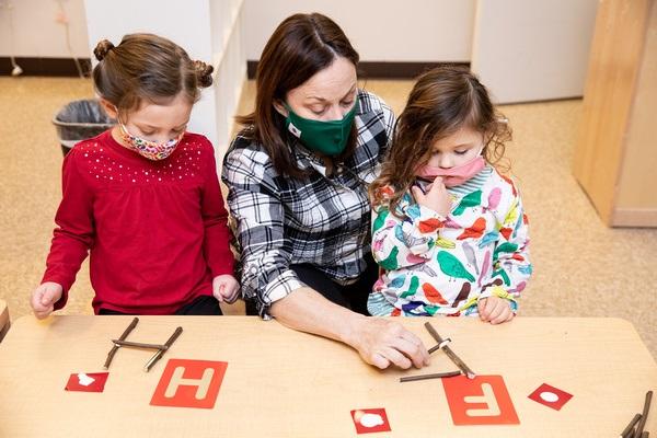 A teacher sits at a table between two children. The H and F stencils are on the table. One child makes an H with sticks. The teacher helps the other child make an F with sticks.