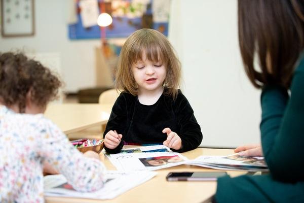 A teacher and two children sit at a table writing with crayons.