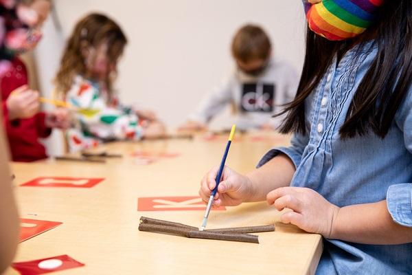 Children sit at a table with letter stencils and stick letters. They trace the stick letters with a paintbrush.
