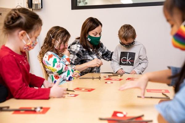 A teacher and children sit at a table looking at and touching letter stencils and stick letters.