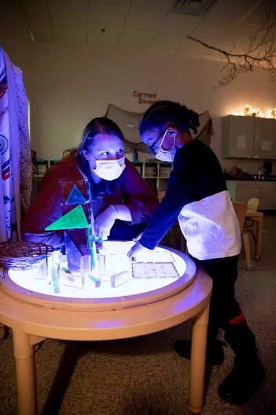 A teacher sits and child stands at a lighted table touching clear shapes.
