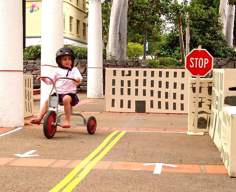 A child rides a tricycle down concrete that has white arrows showing which direction to ride on each side of a double yellow line. A stop sign stands at the end of one side of the pretend road.