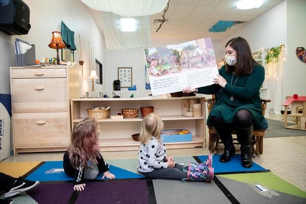 A teacher sits in a chair holding a big book with the pictures and print pointed toward the two children sitting on the carpet. The two children look at the book.